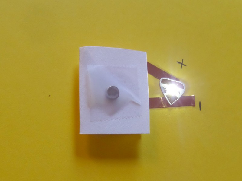 A folded piece of  paper with a round disc magnet taped to the top with Scotch Tape connects to the positive and negative leads of a white Circuit Sticker LED, which is glowing.