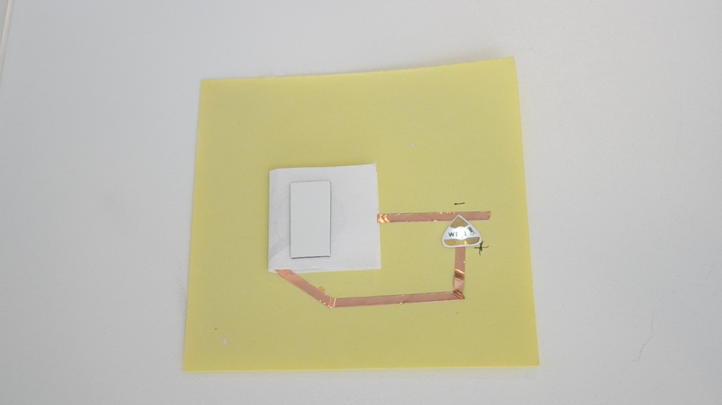 A folded piece of paper is attached to the top center of a yellow piece of paper.  A rectangle of sheet magnet rests on top, suggesting the presence  of a coin battery inside.  To the right, a white Circuit Sticker LED glows.  It connects to the hidden battery with copper tape.