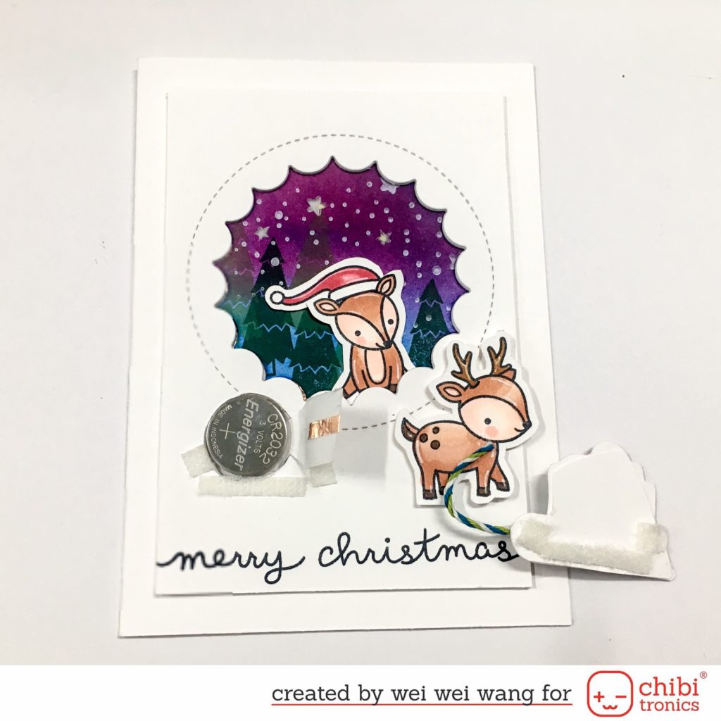 A white card  with a purple medallion in the middle sits upon a white surface.  In the center of the medallion is a squirrel wearing a Santa hat.  To the left of the squirrel is coin cell battery that is touching part of a circuit.  A flap on the right would complete the circuit if it were pressed down upon the battery.  The back side of a paper door, adhered with Velcro tape, rests on the right side of the card.