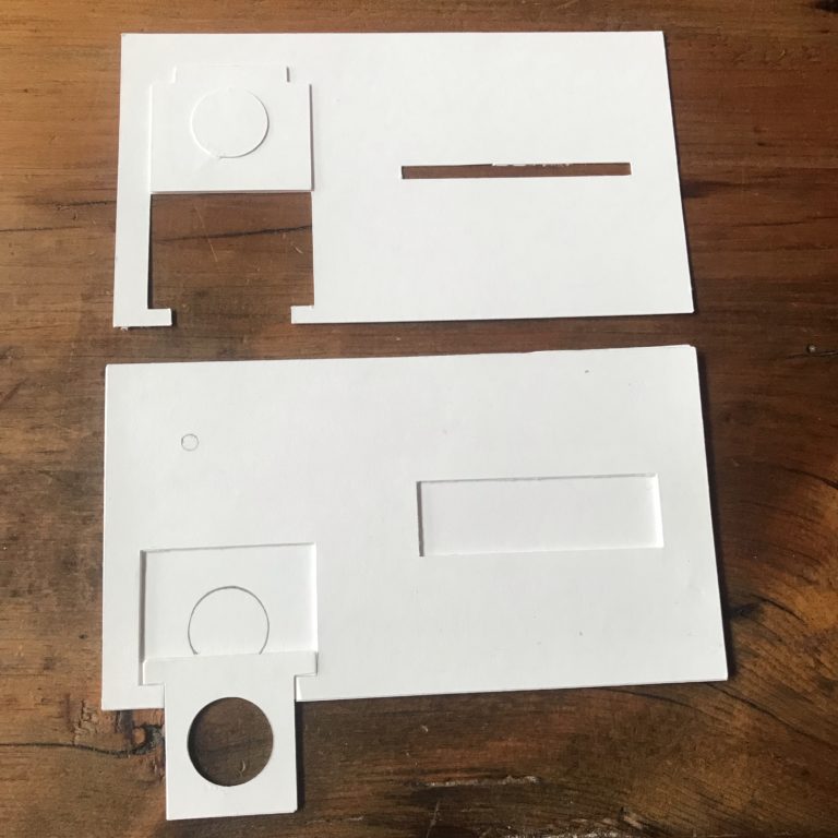 Two plain, white parts of a card rest upon a wooden table.  The top piece has a flap that is designed to rest upon the top of a coin cell battery.  The bottom piece contains a tray made of paper with a hole large enough for a con battery to sit in.