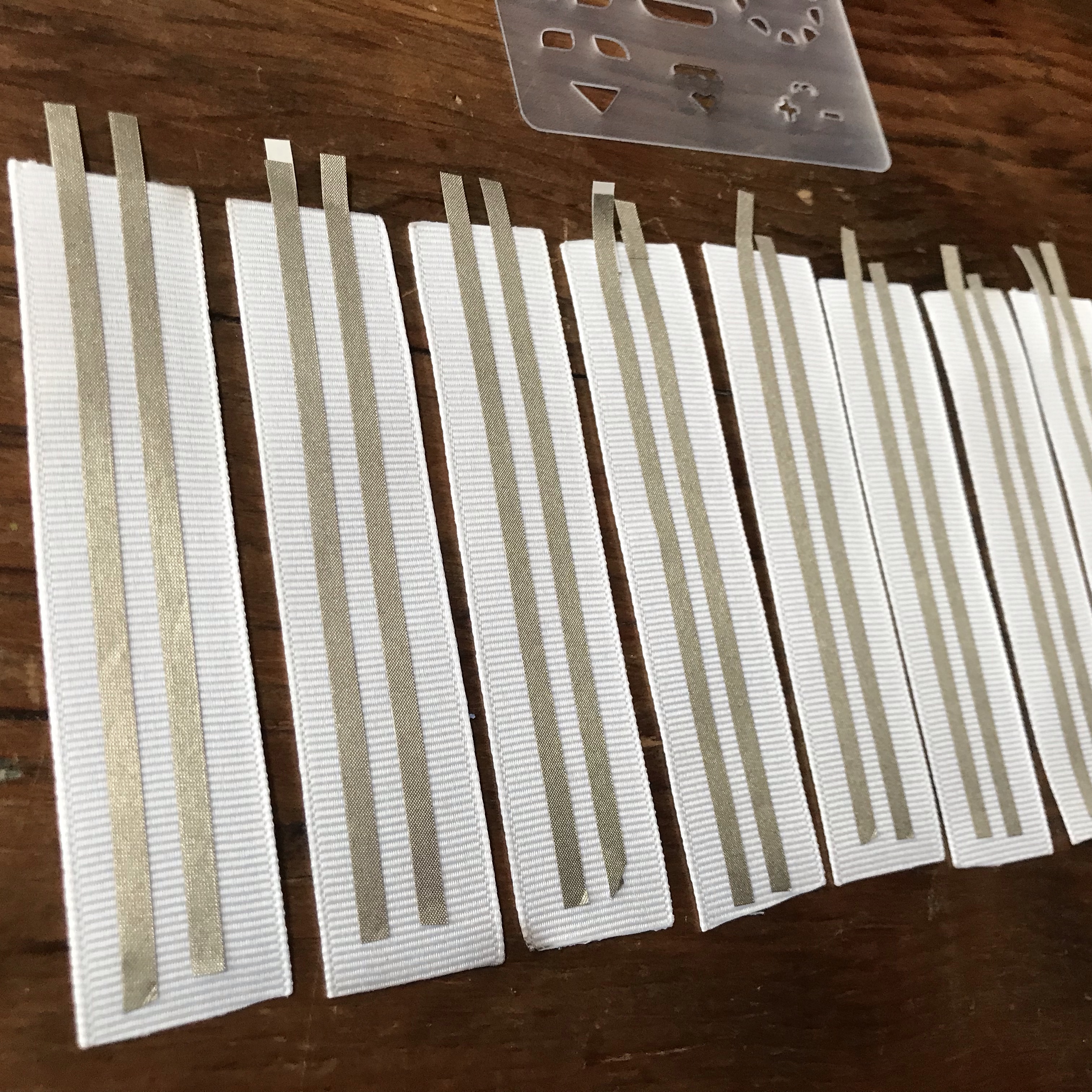 Seven pieces of 1 inch wide X 3" long white ribbon lay side by side upon a wooden table.  Each has two parallel traces of conductive fabric tape adhered in the center, with enough space between the leads for a Sticker LED.