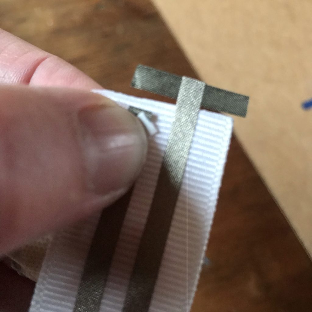 A 1 inch wide piece of ribbon with two parallel leads made of conductive fabric tape.  The negative lead on the right has a perpendicular piece of conductive fabric tape adhered to it with the sticky side up, to allow it to attach to the back, negative lead of a ribbon rail.