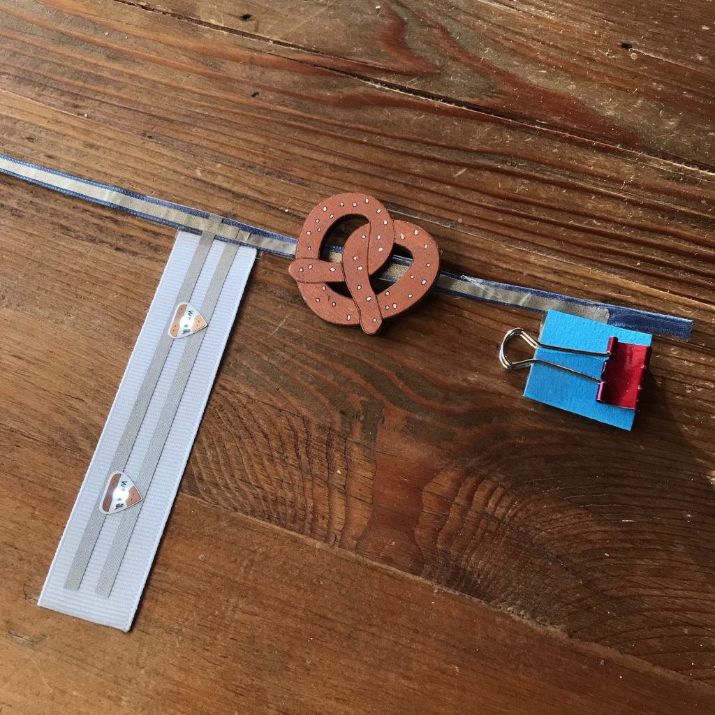 Close up of the positive side of the ribbon rail. A flag with two lit LED stickers is shown, and the magnetic pretzel decoration is shown activating a concealed reed switch to test the circuit. The battery holder (now on the right) is held closed with a small red binder clip.