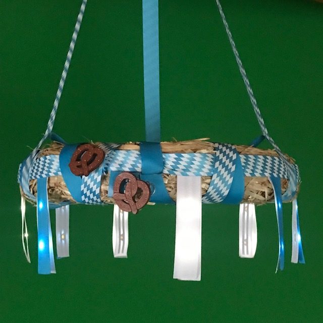 A straw wreath wrapped in blue and white ribbon hangs suspended against a green background.  Two wooden pretzels are attached to the front side of the wreath.