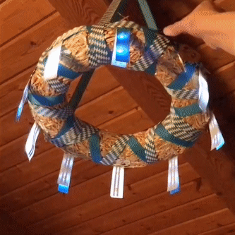 A straw wreath form, wrapped in blue and white ribbon, is suspended from a wooden ceiling.  Nine ribbon "flags" hang from the outer perimeter.  A hand grabs a wooden pretzel on string, a pretzel with a magnet on the back of it, and flips the pretzel over.  The magnetic pretzel causes the LEDs to glow.