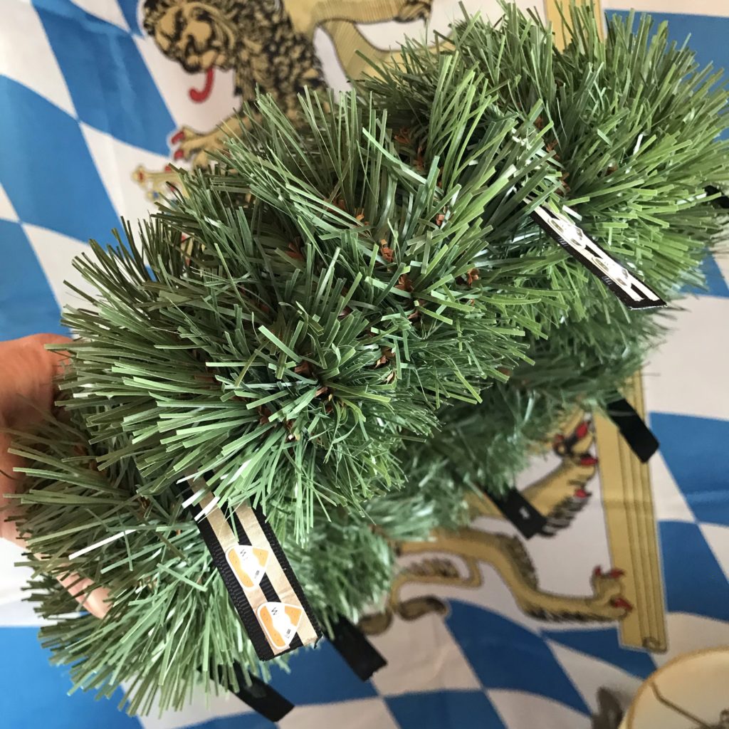 A wreath form wrapped in fake pine tinsel is shown in front of a Bavarian flag.  Glowing ribbon flags extend from the wreath's outer perimeter.  Sticker LEDs bridge the gap between parallel leads running down the center of each flag.  