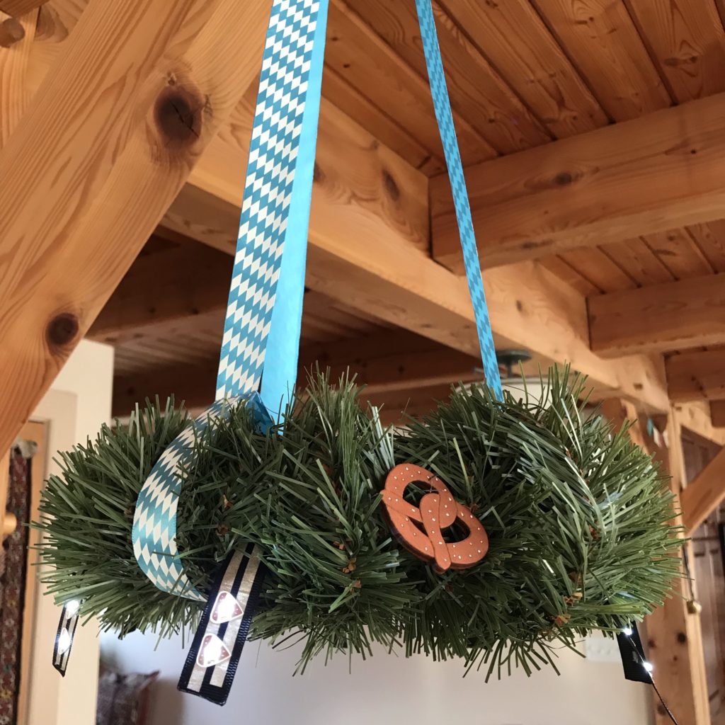 A wreath wrapped in fake pine garland is suspended from a wooden ceiling.  Two glowing LED flags hang down from the outer perimeter of the wreath.  A wooden pretzel decoration is attached to the center of the wreath.