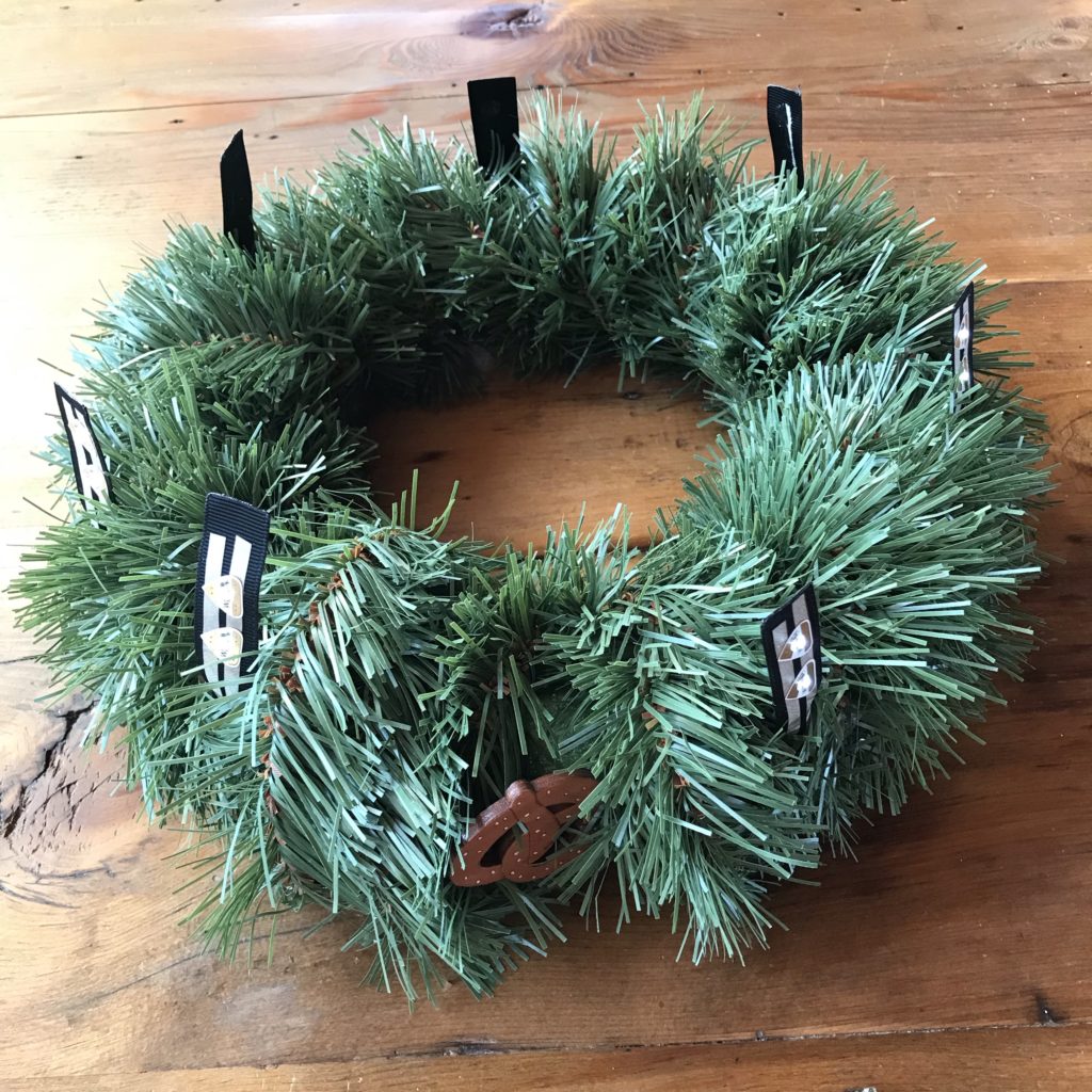 A wreath form completely wrapped in fake pine tinsel lays upon a wood table.  Seven glowing LED flags extend from the outer perimeter of the wreath.  A magnetic pretzel made of wood has been tied to the wreath near a concealed reed switch, to keep it from falling off.