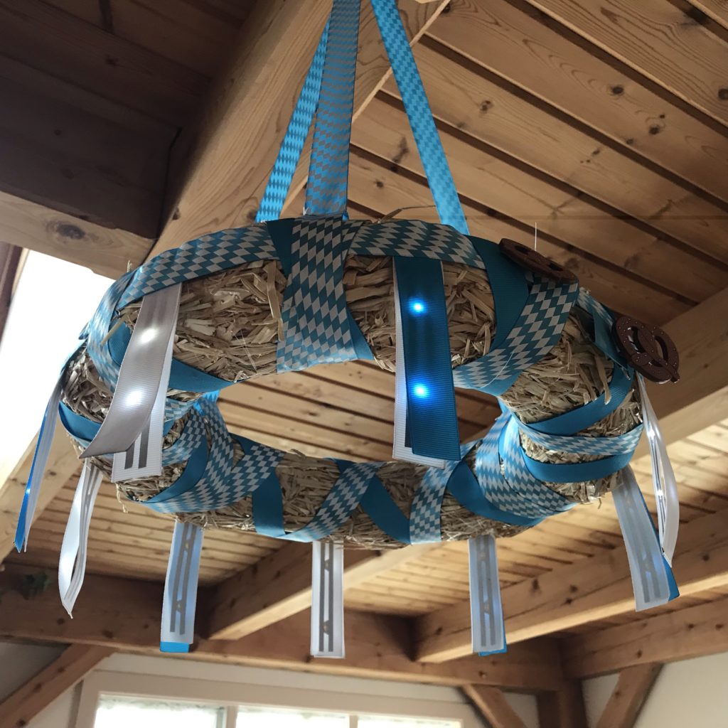 A straw wreath form wrapped in blue and white ribbon is suspended from a wooden ceiling.  Nine ribbon "flags" containing glowing Sticker LEDS hang from the outer perimeter of the wreath.