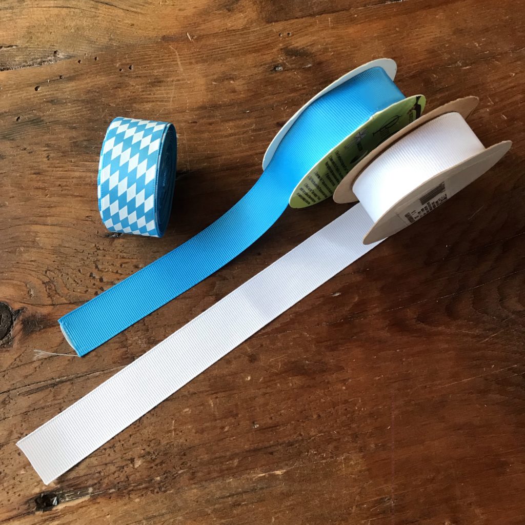 Three rolls of 1" wide blue and white ribbon sit side by side on a wooden table.  The first one has a blue and white diamond motif.