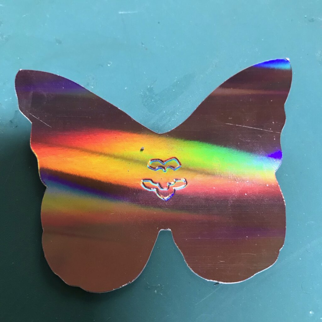 An iridescent, butterfly-shaped, piece of paper rests upon a green cutting mat.  The center of the butterfly has been embossed with a footprint for a Circuit Sticker LED (with its negative end oriented at the top and its positive end oriented toward the bottom). 