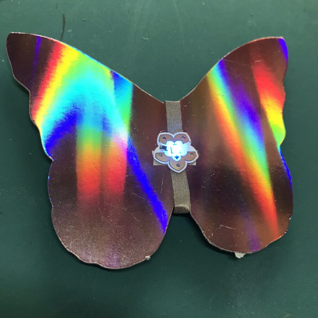 A flower-shaped Animating LED has been adhered on top of the positive and negative leads created upon an iridescent, butterfly-shaped piece of paper.  The narrow side of the LED sticker with two petals is in contact with the negative lead (top).  The broader side of the sticker with three petals in in contact with the positive lead (bottom).