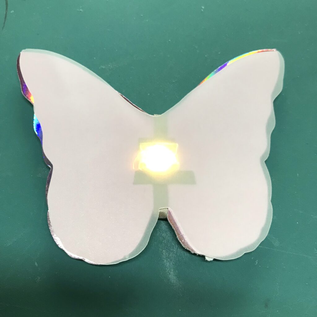 A butterfly-shaped piece of translucent vellum with a hole in the center has been placed over the iridescent butterfly piece containing an LED. The pieces are connected by a small glue dot over the hole.