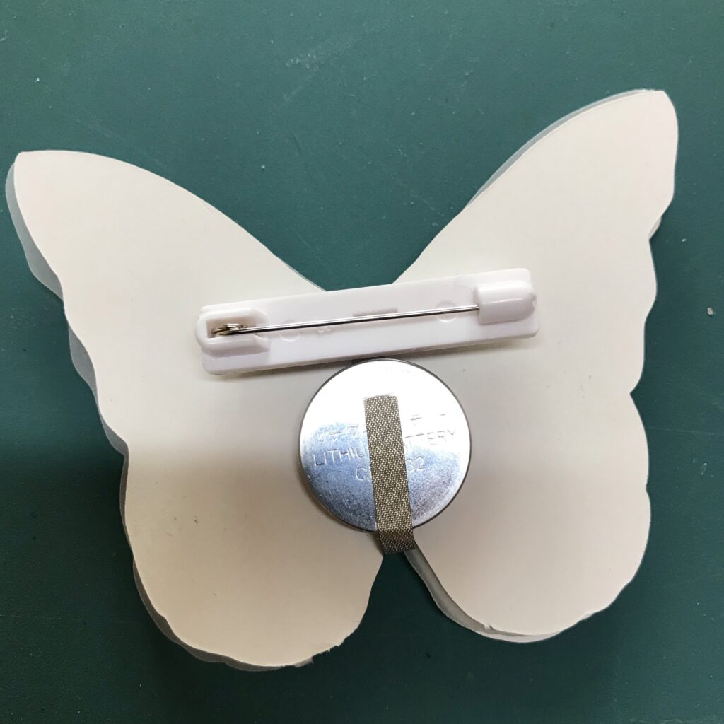 A white plastic, adhesive-backed, pin has been adhered to the back side of a butterfly-shaped circuit that has a coin cell battery connected to it.