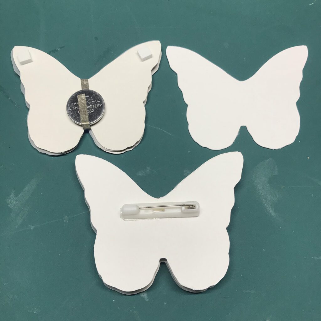 shaped piece of paper about to be placed on top of the foam squares to make room for the addition of the plastic pin back.  The bottom butterfly piece has an extra piece of paper attached to its wing tips and a pin back centered on it.