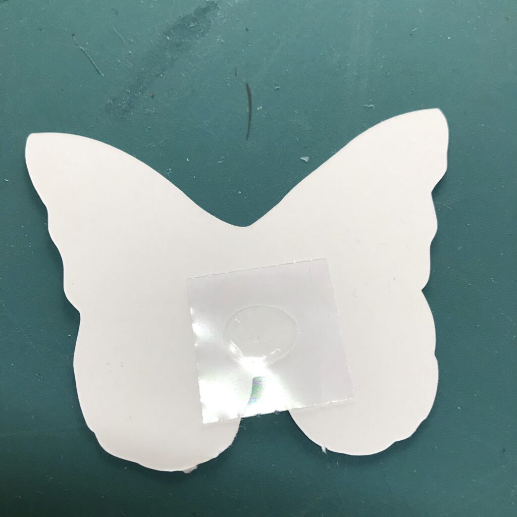 The white, non-iridescent back side of a butterfly-shaped piece of paper contains a glue dot .  The glue dot has been placed slightly below the center of the butterfly piece and it still has on its clear, plastic backing.