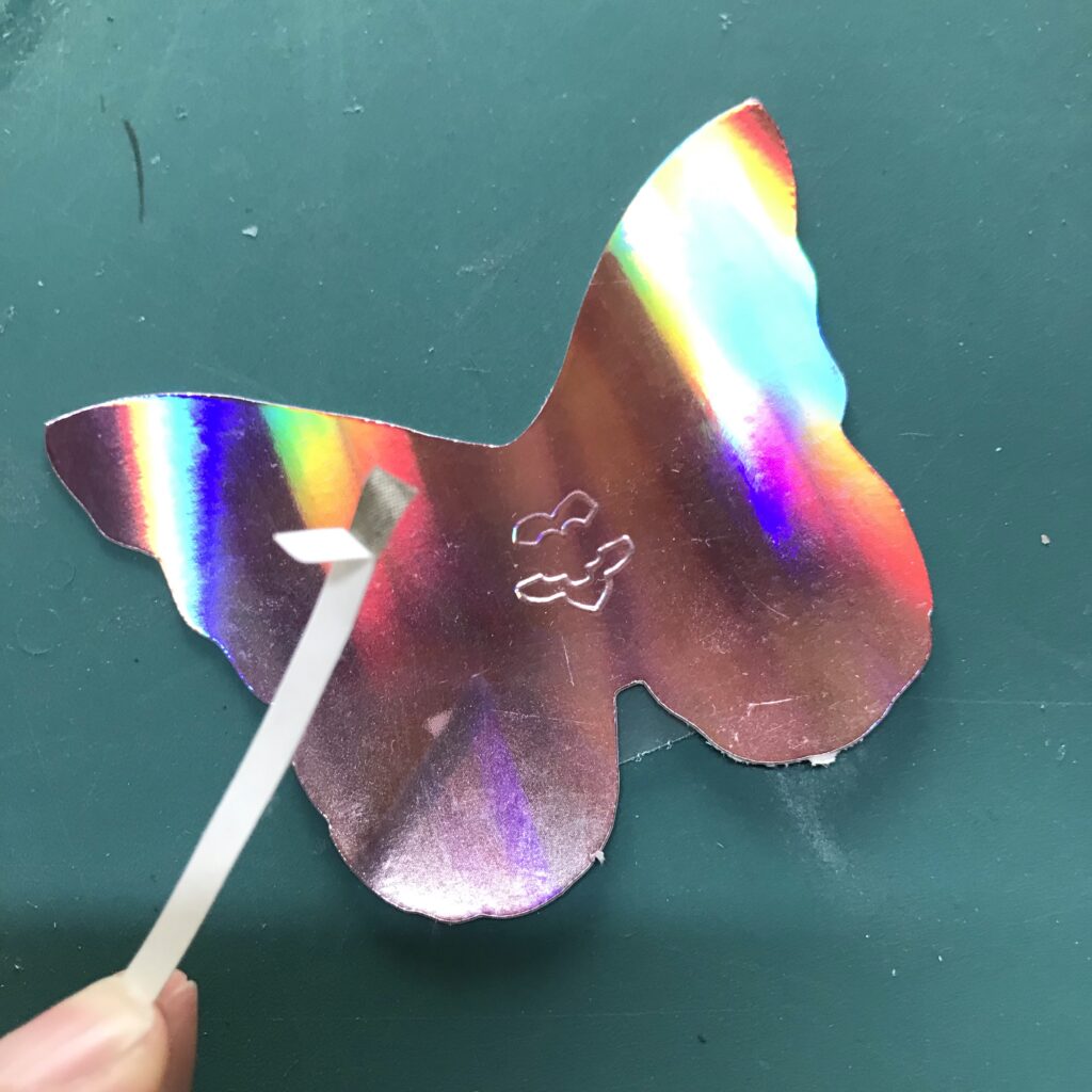 A piece of conductive fabric tape has been prepped by removing part of the protective backing from the top 1/4 inch.  An iridescent butterfly piece with an embossed flower, denoting the footprint for an Animating LED, rests upon a green craft mat.