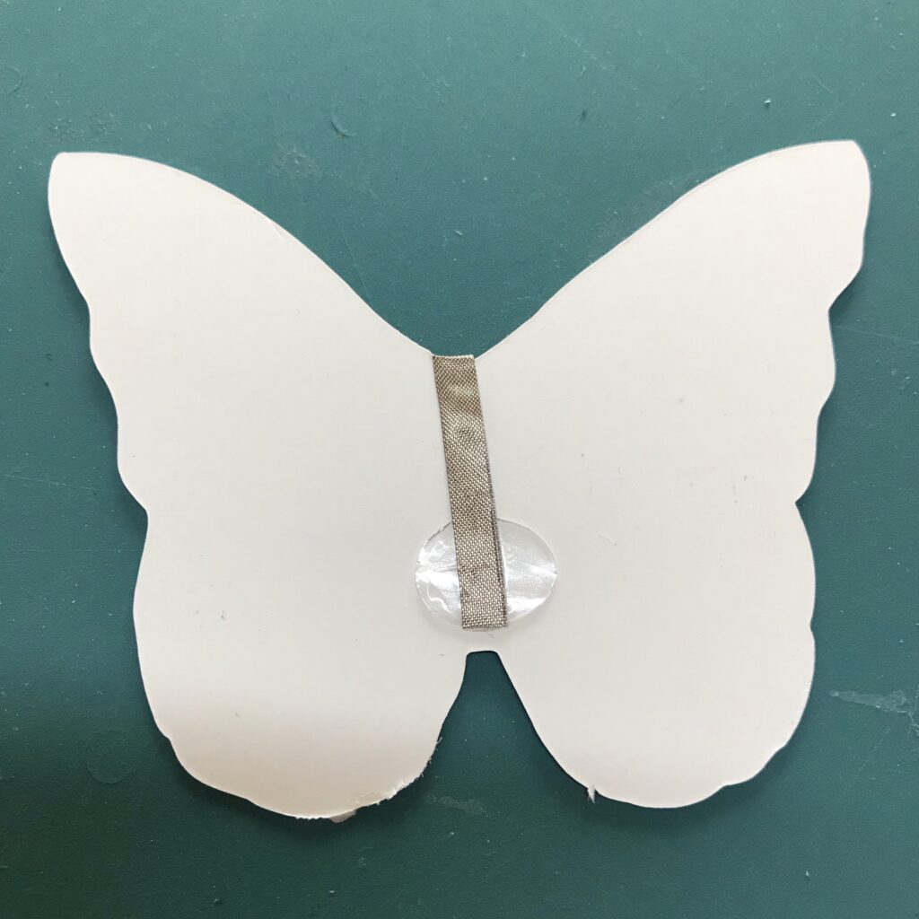 The white, back side of the butterfly-shaped piece of paper contains a glue dot with its plastic backing removed.  A piece conductive fabric tape is overlapping the glue dot and extending (hidden from view) around to the back (iridescent side) of the butterfly piece.