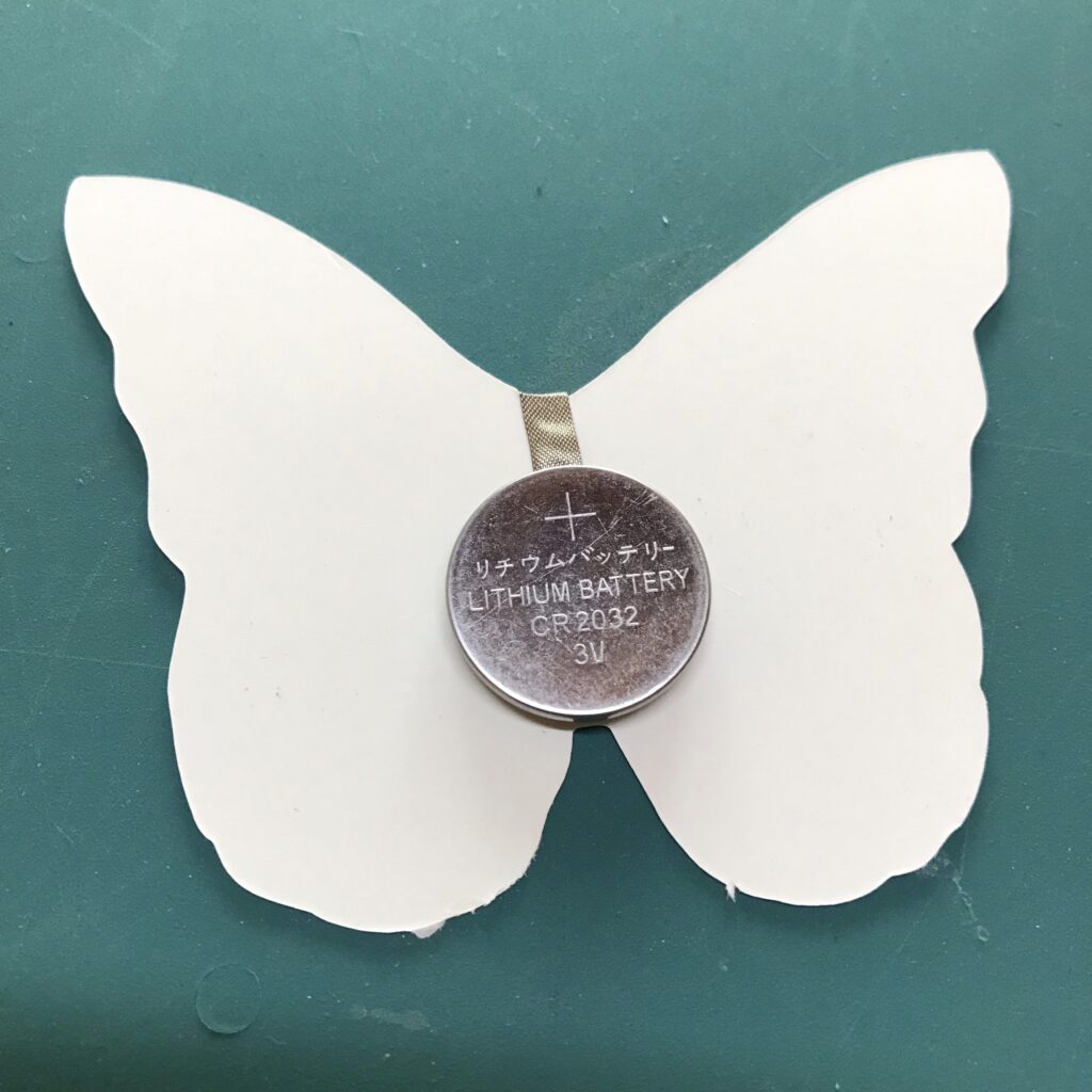 The white, back side of the butterfly-shaped piece of paper contains a coin cell battery with the positive side facing up, denoted with a + sign. A piece conductive fabric tape is under the battery, extending (hidden from view) around to the back (iridescent side) of the butterfly piece.