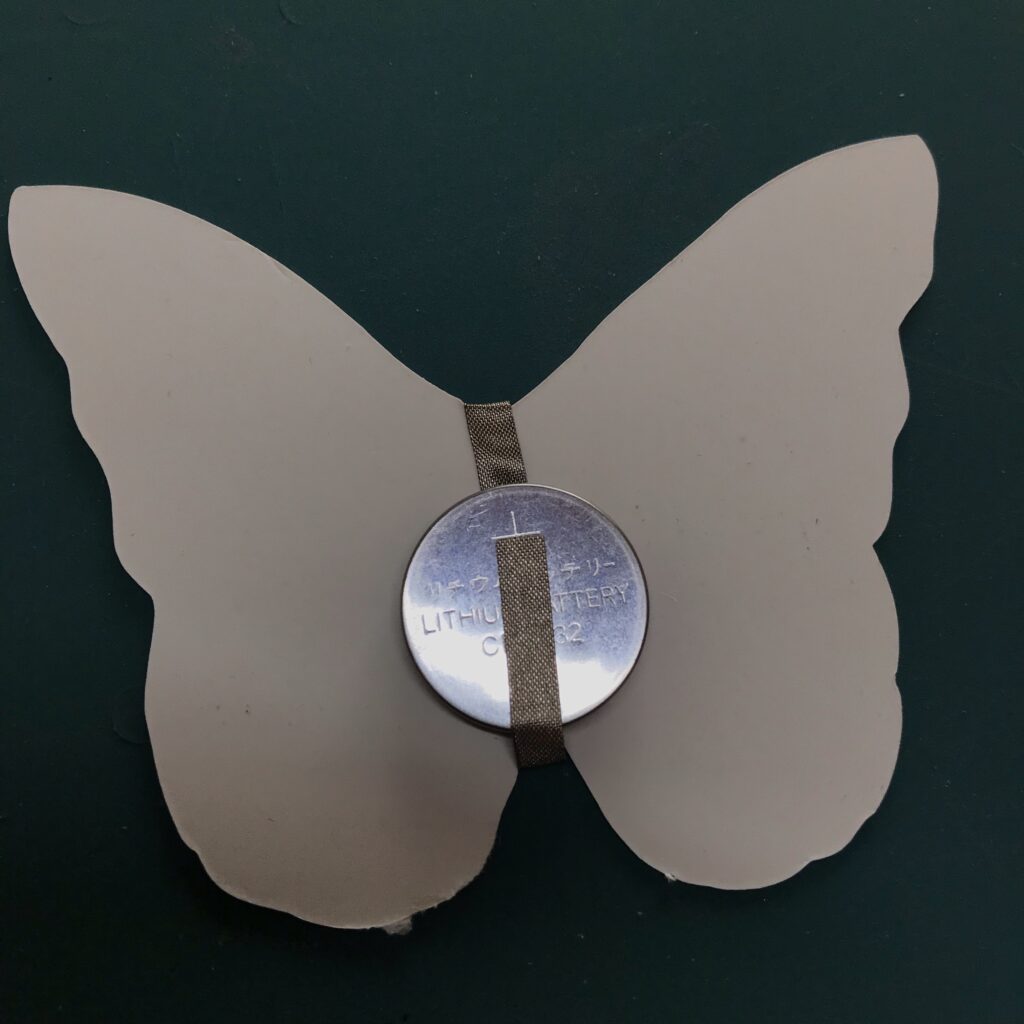 The white, back side of the butterfly-shaped piece of paper contains a coin cell battery with the positive side facing up, denoted with a + sign.  A piece conductive fabric tape is under the battery, extending (hidden from view) around to the back (iridescent side) of the butterfly piece.  A second piece of conductive fabric tape has been applied to the top side of the battery (from the front side, hidden from view).  The piece of tape that is on top of the battery does not touch the piece of tape that is under the battery.