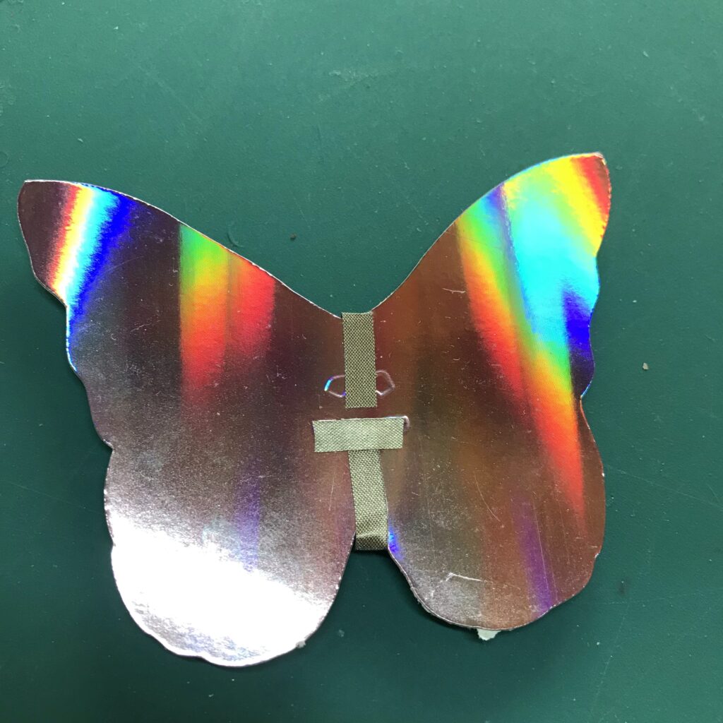 The front, iridescent side of a butterfly shape, with an embossed flower in the center to denote the placement of an Animating LED, has negative and positive leads starting on the front side and wrapping around to the back side (hidden from view).  A small piece of conductive fabric tape has been applied perpendicular to the positive lead, without touching the negative one.