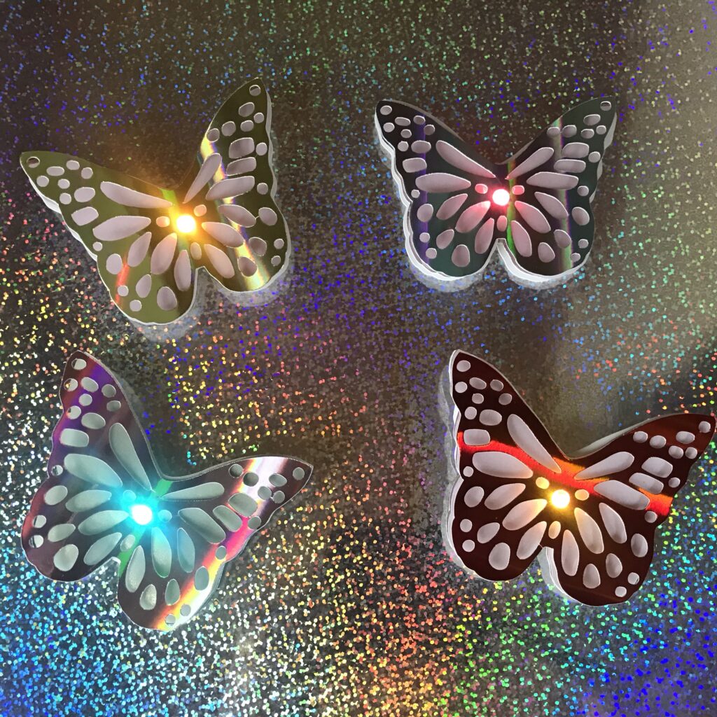 Four lacy, iridescent butterfly pins with different LEDs glowing from the centers of their bodies rest upon a piece of silver holographic paper.