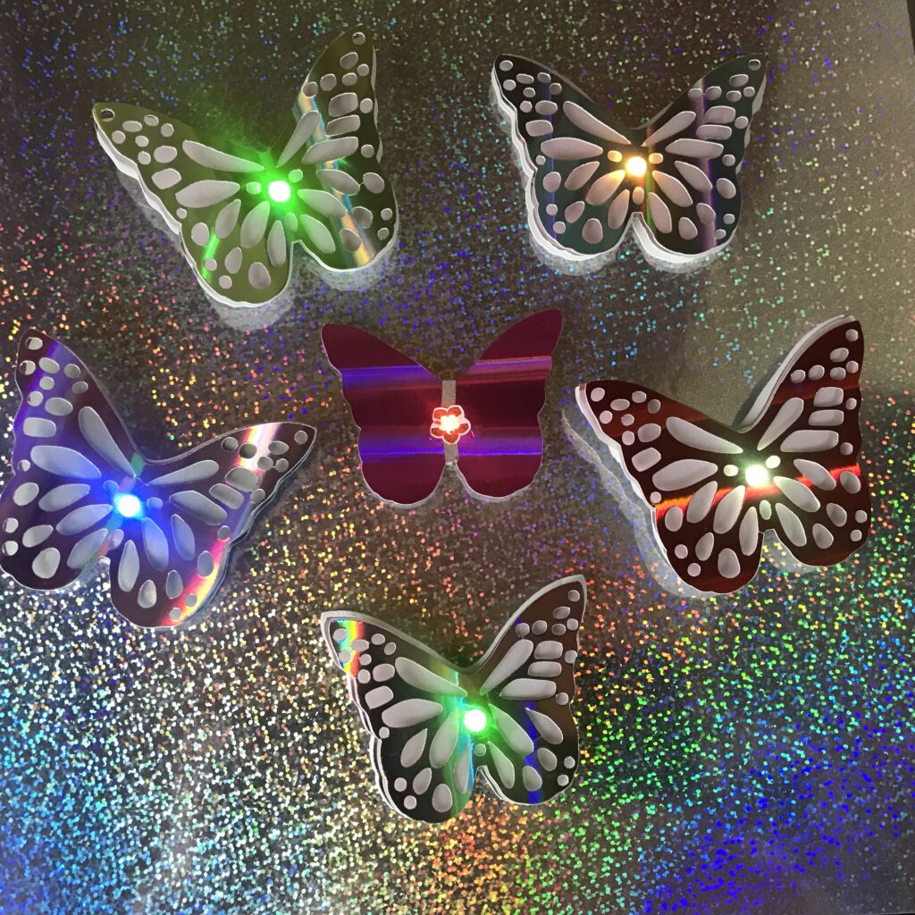 Six butterfly shaped pins with glowing LEDs in the centers of their bodies are placed on top of a holographic background.  A red butterfly in the center reveals a flower-shaped LED placed on top of two pieces of conductive fabric tape, while the remaining five have decorative layers concealing the circuit.