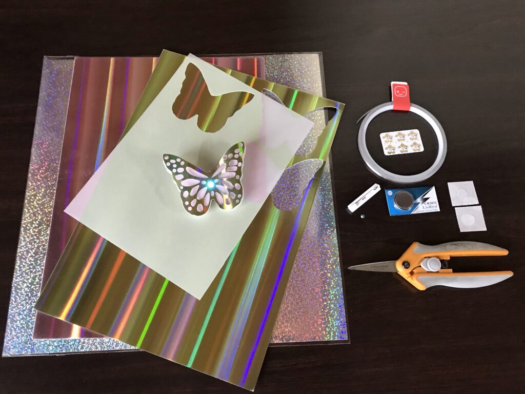 the pile, along with a completed butterfly pin with a glowing blue LED in the center of its body.  To the right are a roll of conductive fabric tape, six flower-shaped animating LEDs, a plastic pin back, a coin cell battery in its packaging, two glue dots with a protective backing a transparent gemstone, and a pair of scissors. 