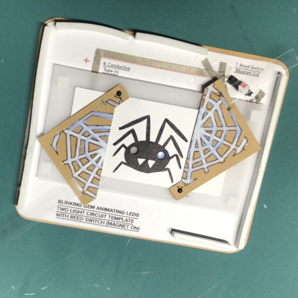 A webbed set of diagonal doors, made from paper, reveals a black spider with a glowing white eye at the opening.  The doors are resting on top of the completed circuit on the back panel of a card.