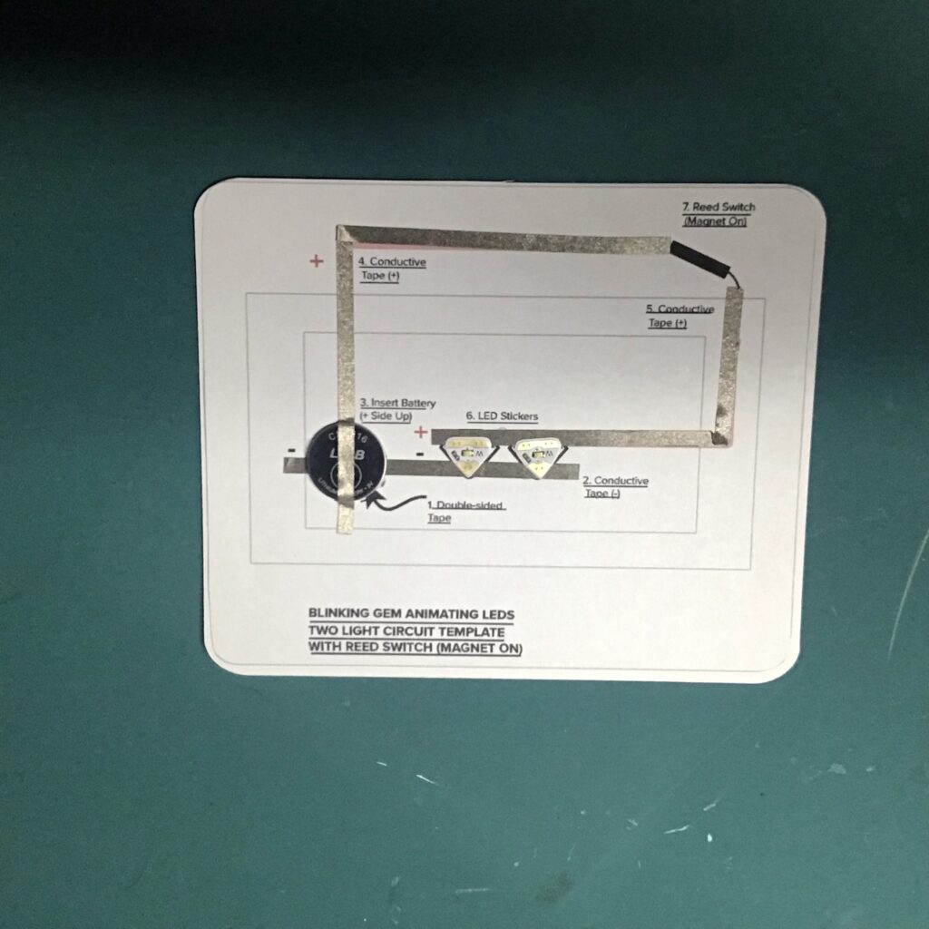 Two Circuit Sticker LEDs have been adhered to the center of a circuit diagram., resting on top of conductive fabric traces.