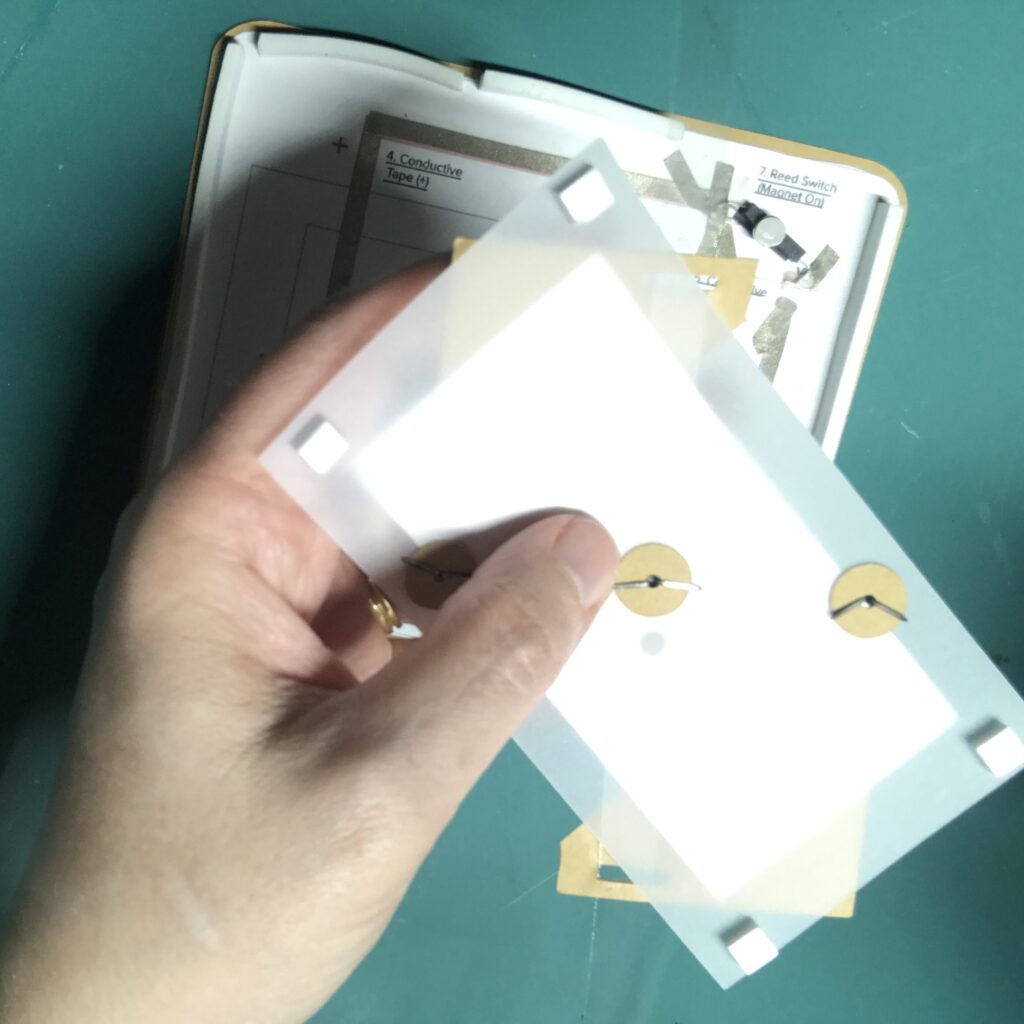 The back side of a rectangular piece of vellum has three paper washers held in place with metal brads.  Each of the four corners has a square of foam tape  attached to it.