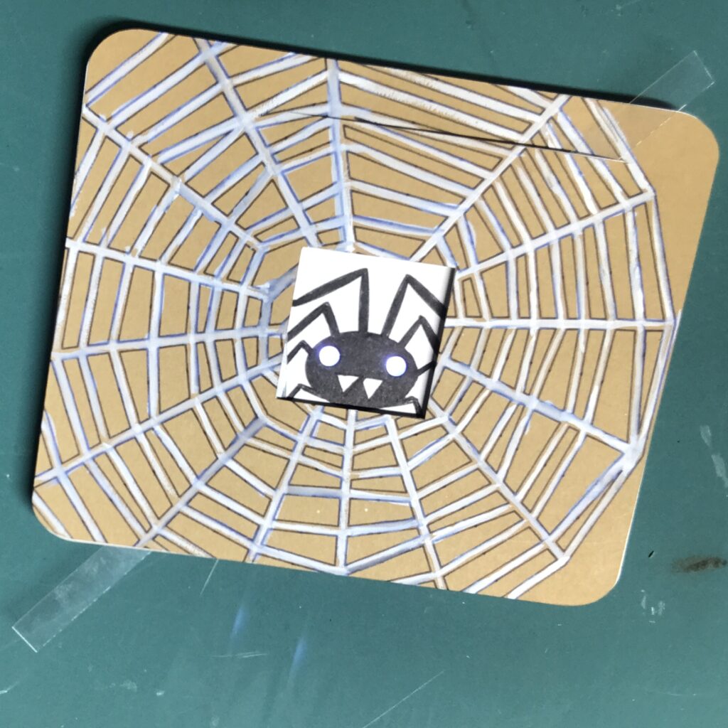 A clear strip of acetate has been inserted through the slot in a rectangular card front.  A black spider against a white background has two glowing, white LED eyes.