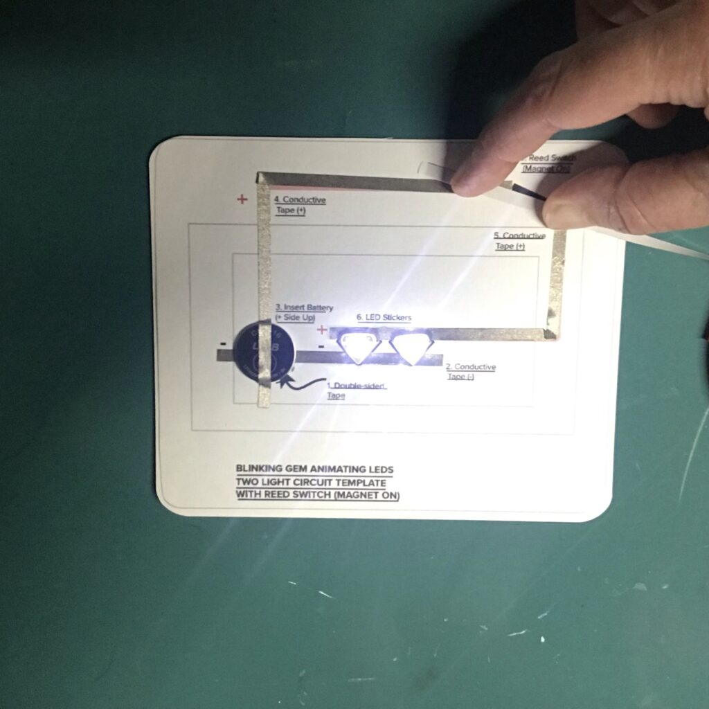 A hand is holding a piece of conductive fabric tape over a gap in the positive lead of a circuit in order to close the circuit. Two Circuit Sticker LEDs are glowing.