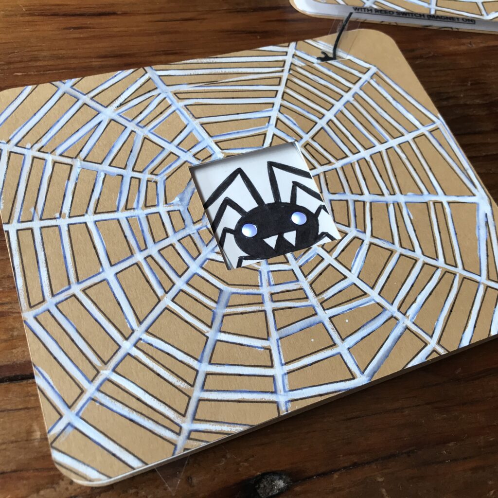 A brown, rectangular card with a white spider web rests upon a wooden table. In the center of the card is a black spider against a white background with white glowing LED eyes.