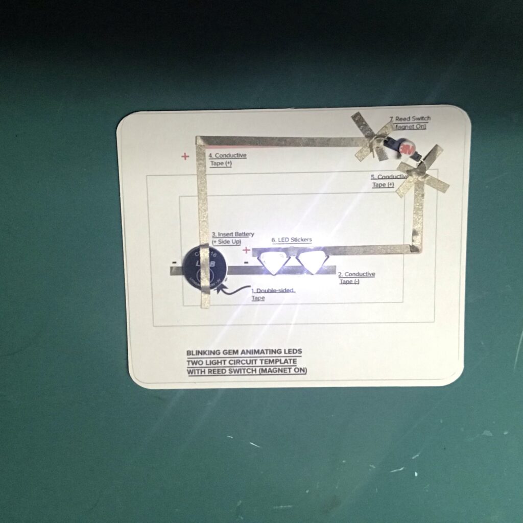 A reed switch with curved legs has been adhered to the top right corner of a circuit template with pieces of conductive tape that resemble X's.  A battery rests upon the reed switch, causing two white Sticker LEDs to light up.