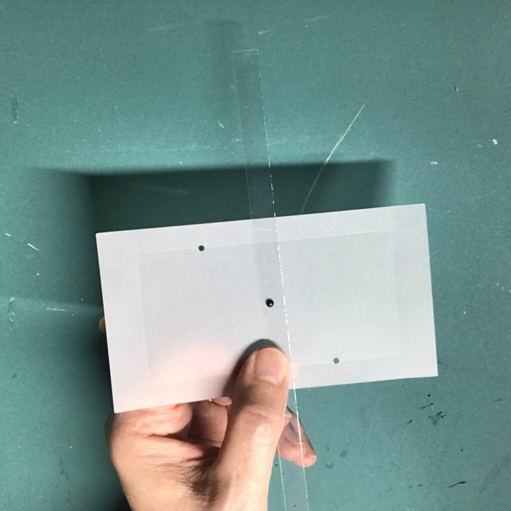 A piece of white vellum with three diagonal holes, rests in a hand. A narrow strip of clear acetate is centered over the top of it.