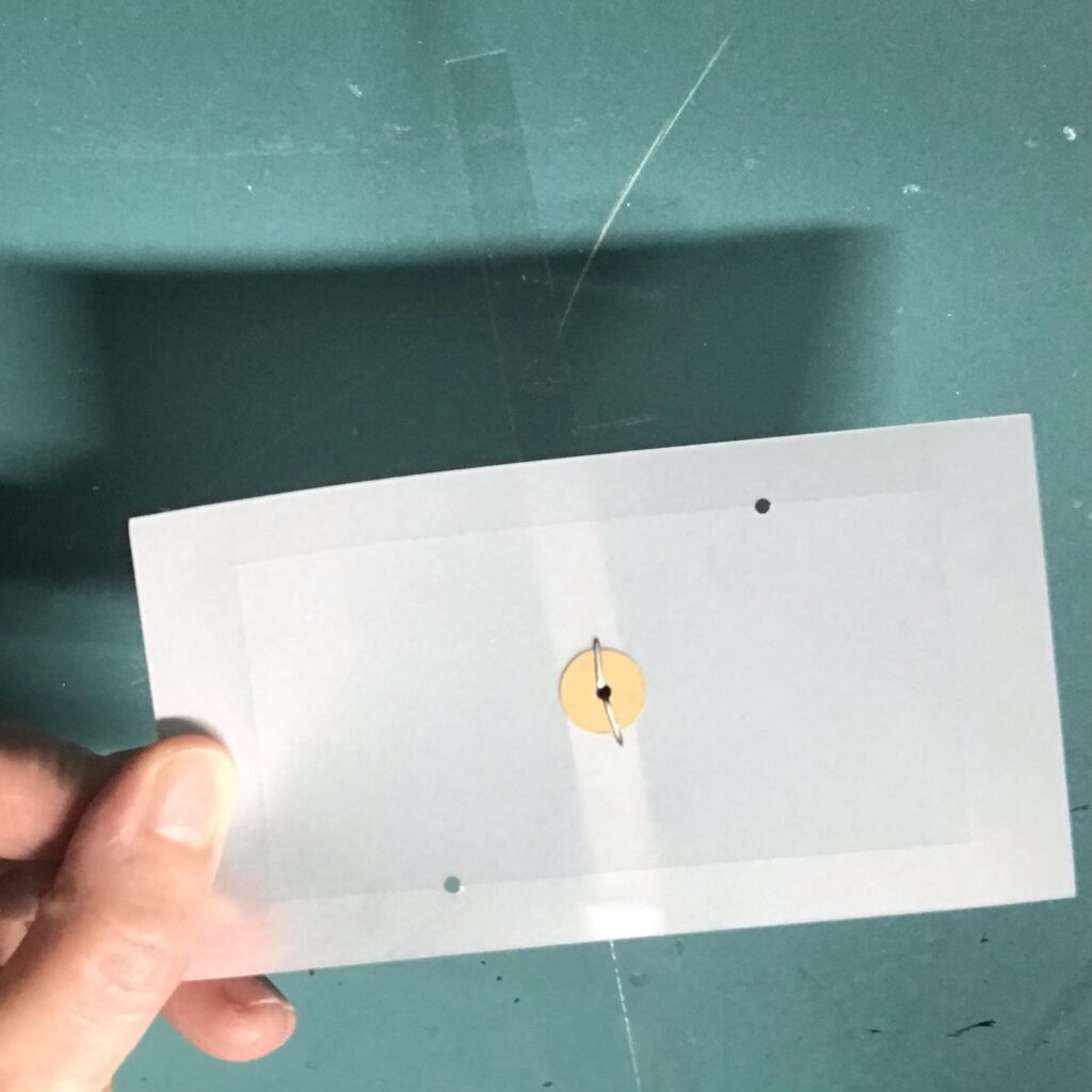 The back view of a piece of vellum with three diagonal holes.  A clear acetate strip has been secured to the opposite side with a metal brad and secured to the back with a paper washer.