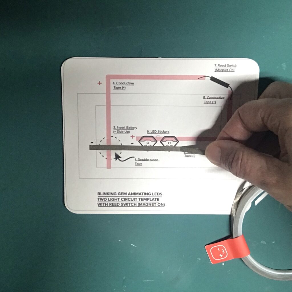 A piece of conductive fabric tape is being applied in a straight line, perpendicular to a round footprint denoting where a battery will be placed.