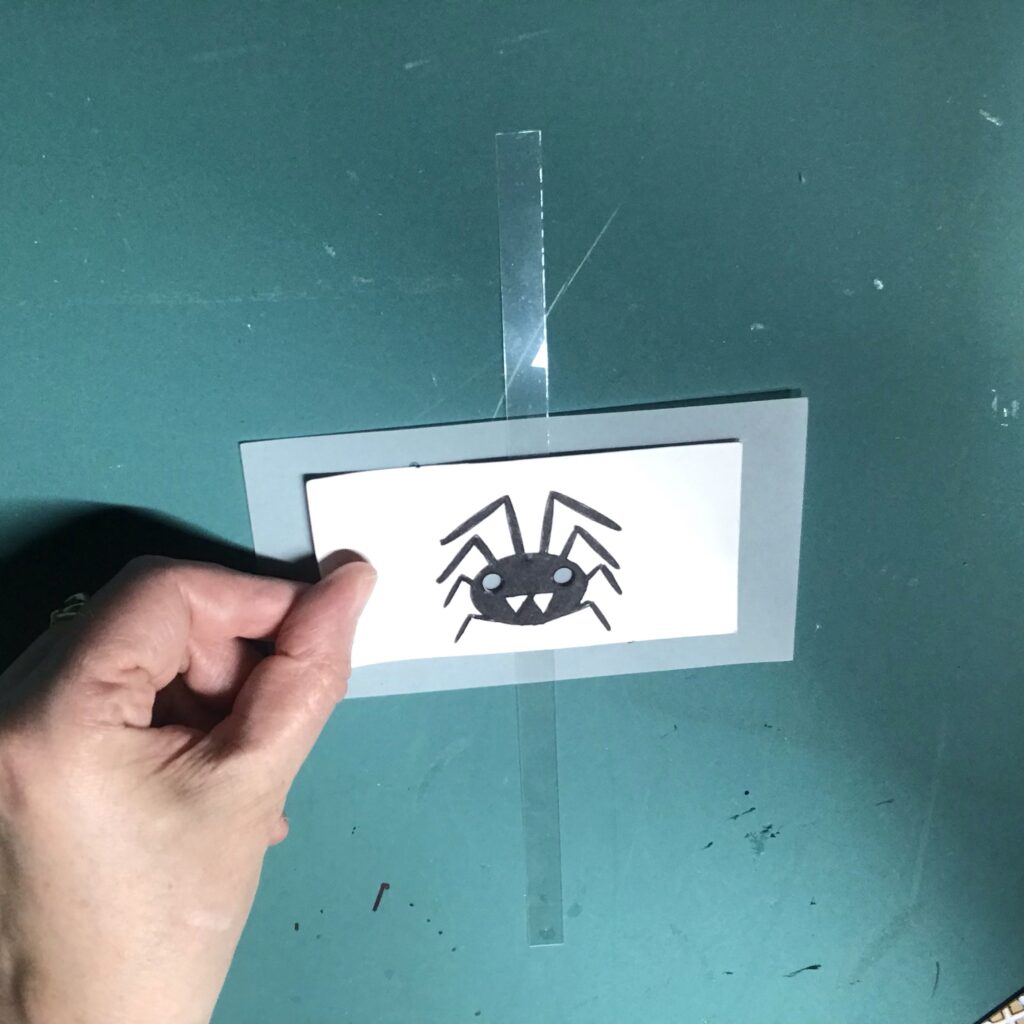A piece of artwork with a black spider on it rests in the center of a white piece of vellum.
