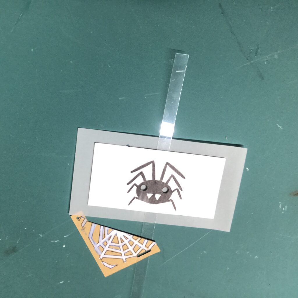A piece of artwork with a black spider on it rests in the center of a white piece of vellum.  A clear strip of acetate runs down the center of it and through a slot on a door that has been cut diagonally.