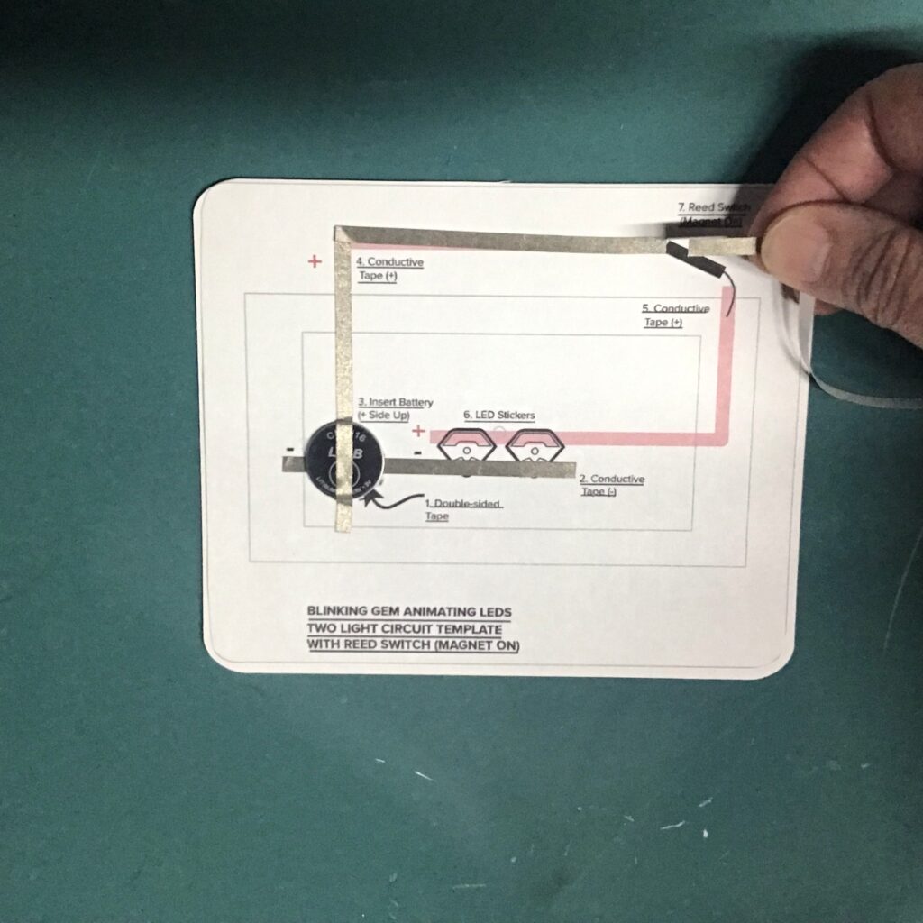 A piece of conductive fabric tape is being applied in the shape of a 90 degree corner, overlapping a CR2016 battery and ending in the top right corner of a circuit diagram.