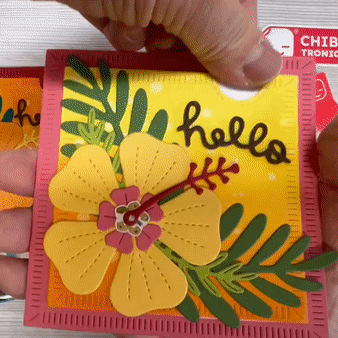 A hand pulls a tab from the top right corner of a square card, decorated with a yellow hibiscus flower. In the center of the flower, a flower-shaped, rainbow fade animating LED changes from red to white to green.
