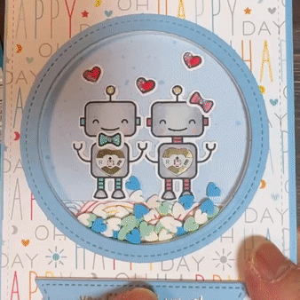 A hand holds a square card with a circular opening that contains two smiling robots. Each robot has a red fade, heart-shaped animating LED in the center of its chest.