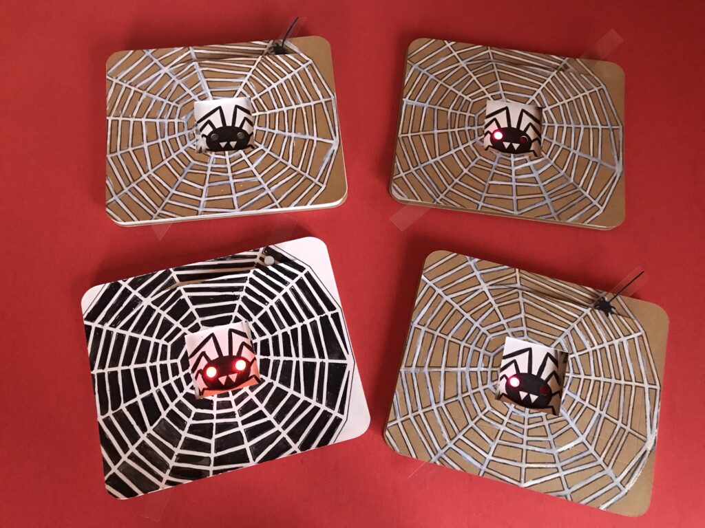 Four Spooky Spider Diagonal Dissolve Cards rest upon a red background. Each card contains a black spider against a white background, in the center.  Each card front has been decorated with a spider web design.  One spider's eyes are black, two have a light shining through a single left eye, and one has both eyes lit.