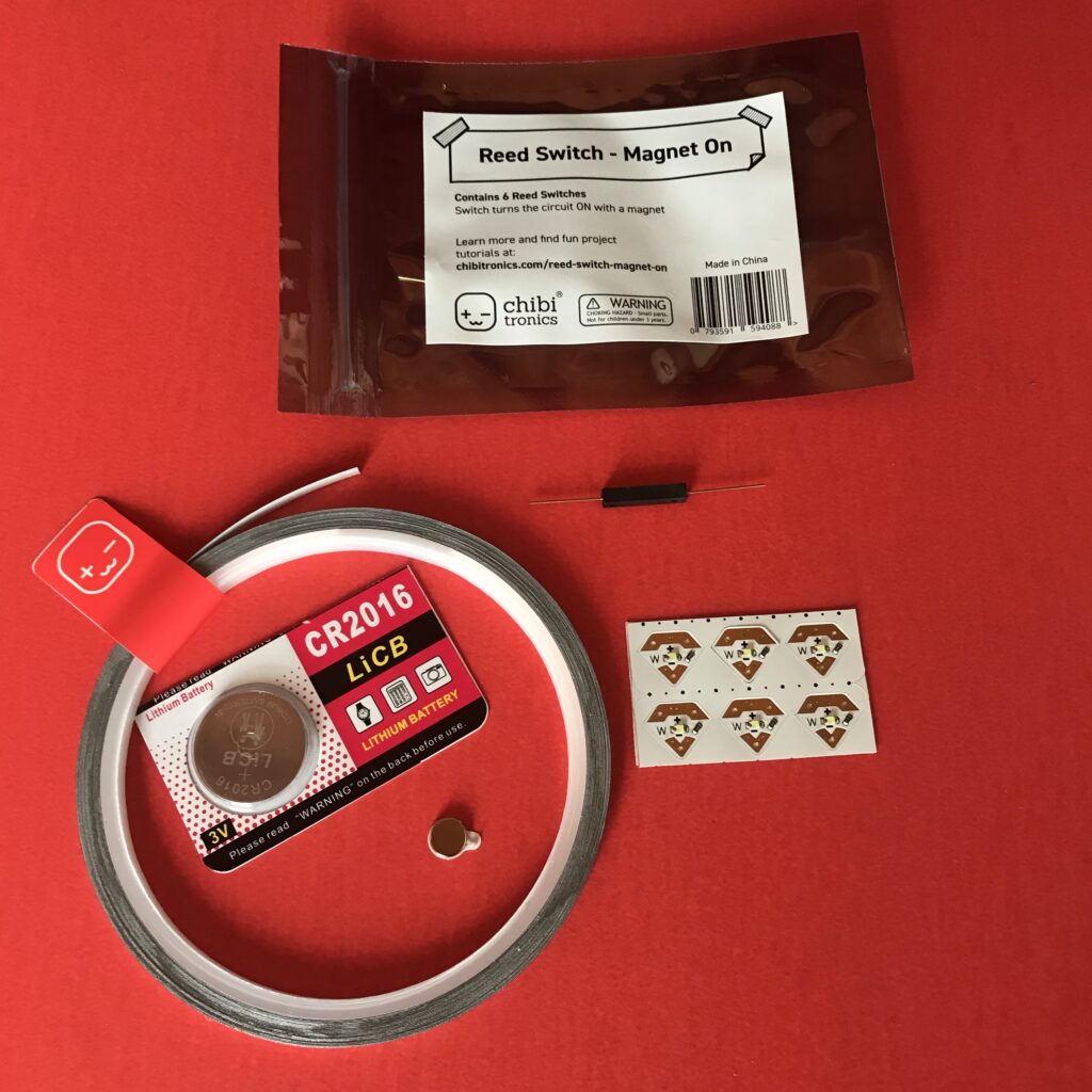 Electrical components needed for making a diagonal dissolve card are placed against a red background: reed switch, CR2016 battery, magnet, conductive fabric tape, Circuit Sticker LEDs