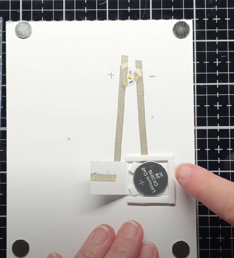 The inside of a white cardstock card panel has four round disk magnets attached in each corner. In the center of the card, a coin cell battery sits on top of a piece of conductive fabric tape that connects with the negative, pointy end of a Circuit Sticker LED.  A paper flap to the right of the battery has another piece of conductive fabric tape wrapped around it and connecting to the broad, positive side of the LED.  Three pieces of foam tape in the same of a three-sided square encase the battery.