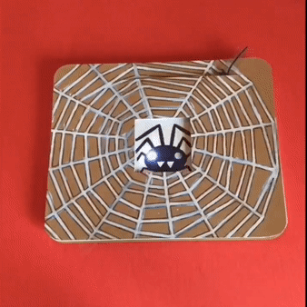 Animated GIF depicting a hand moving a lever on a brown, Spooky Spider Diagonal Dissolve card, which causes a paper door (decorated with a white spider web) to open, revealing a black spider against a white background, with blinkin, white LED eyes.