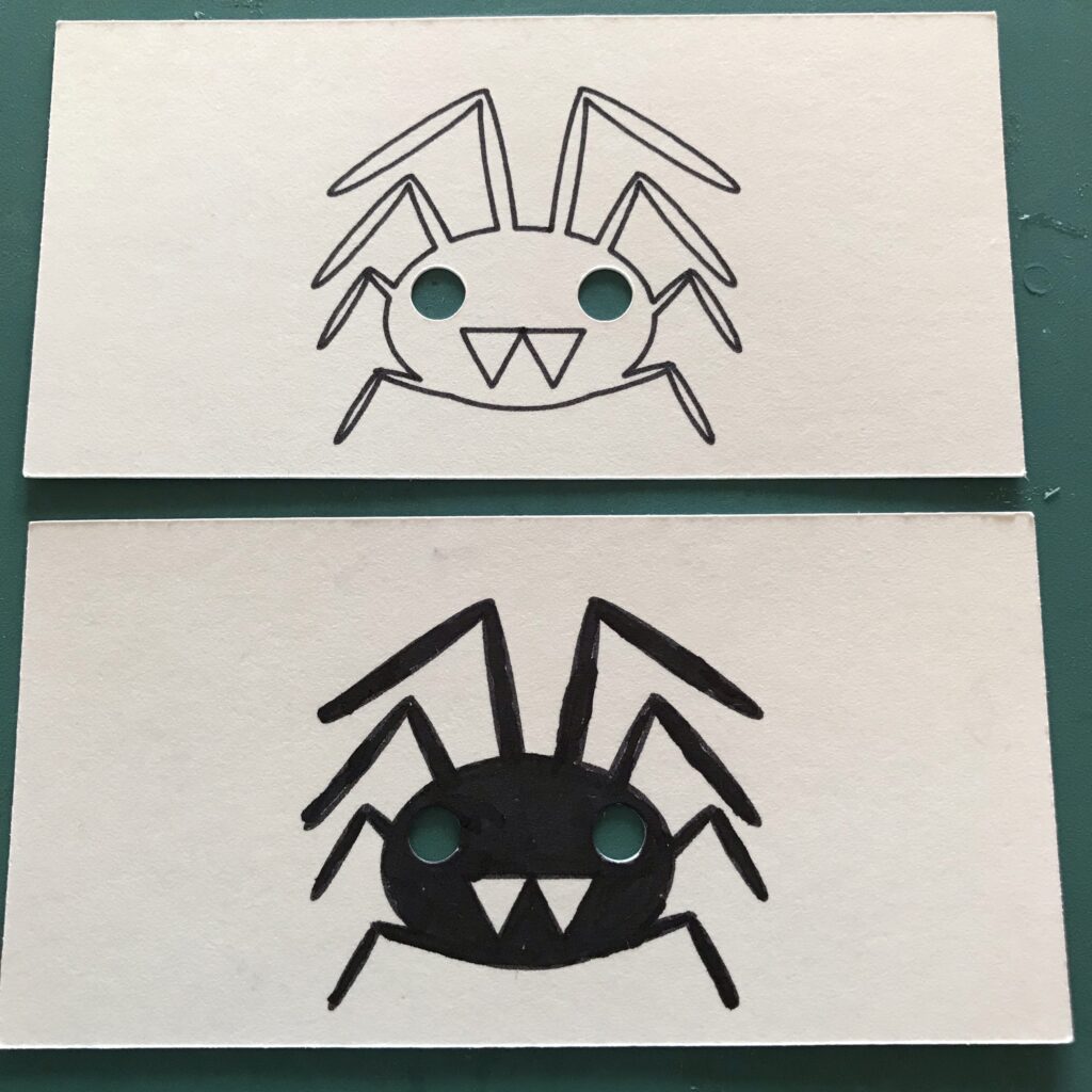 To show comparison, a piece of artwork with an outline of a spider is stacked on top of another piece of artwork that has been colored with a black marker