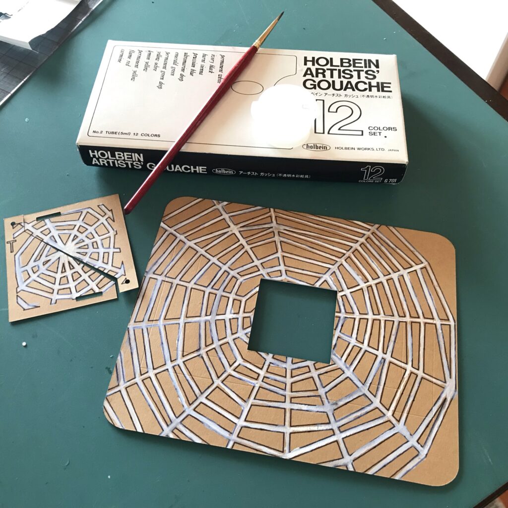 A set of paper doors that have been separated with a diagonal cut sits next to a rectangular card bard. Both pieces are decorated with white spiderwebs.  A box of gouache paints and a brush rests above the painted pieces.