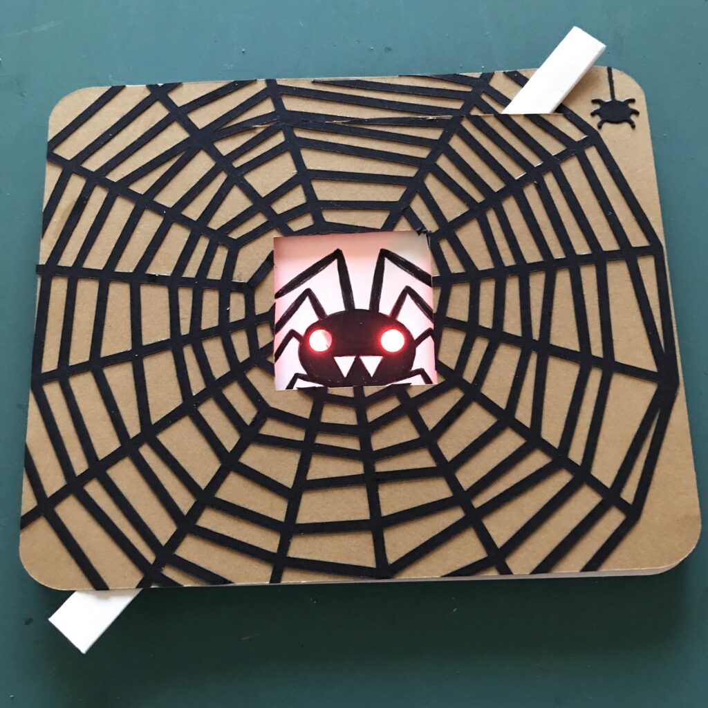 A brown, rectangular card with a black paper spider web glued to the font, rests upon a green background. In the center of the card is a black spider with red glowing eyes and white fangs. A white, paper lever is positioned at an angle inside of the card.